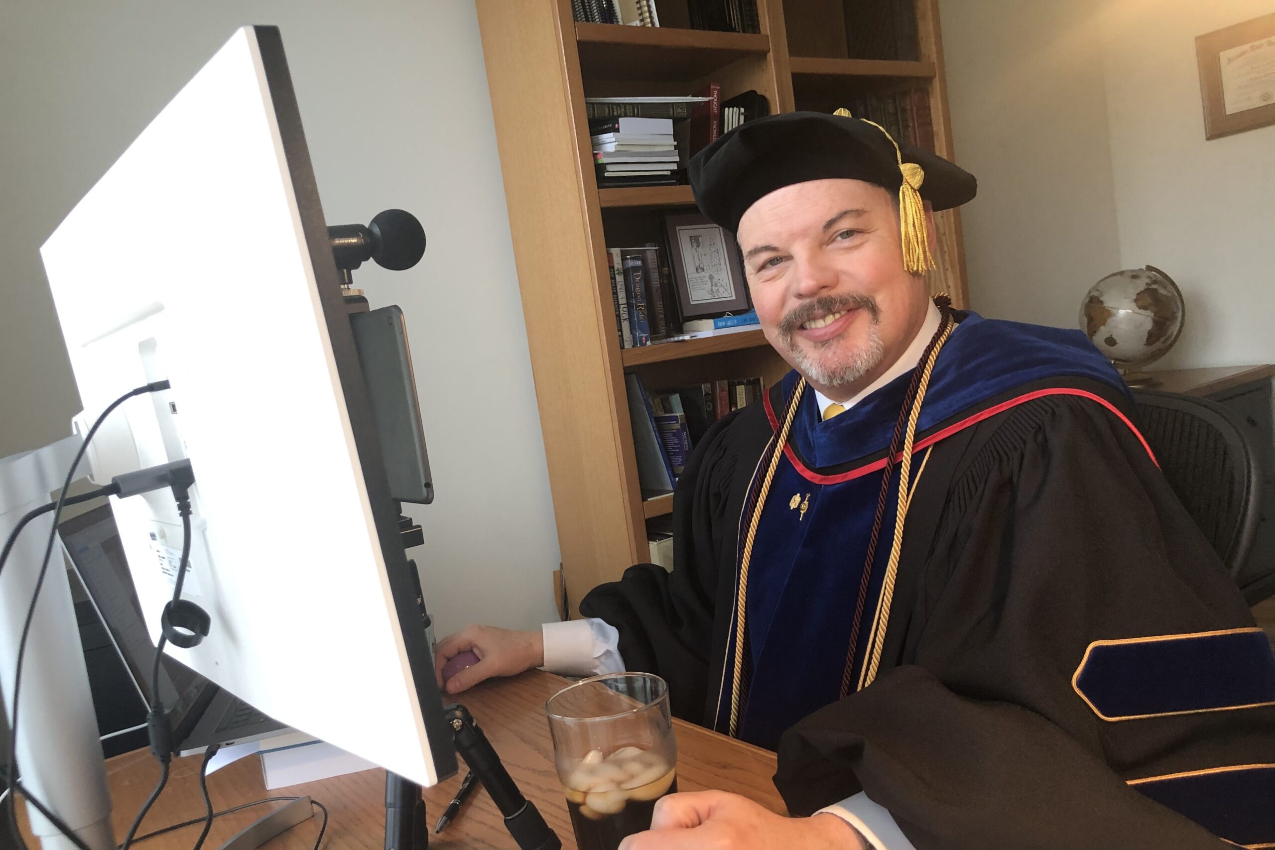 Vernon, a distinguished, middle-age man with a salt and pepper goatee in college graduation robes sitting in front of his office computer.